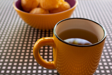 Uma caneca de chá com uma vasilha de pães de queijo sobre uma mesa com forro xadrez de preto e branco.