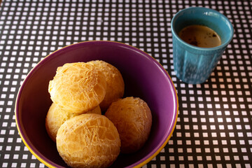 Uma vasilha com pães de queijo e uma caneca com cappuccino sobre a mesa com um forro xadrez de preto e branco.
