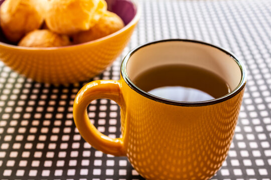 Uma caneca de chá com uma vasilha de pães de queijo sobre uma mesa com forro xadrez de preto e branco.