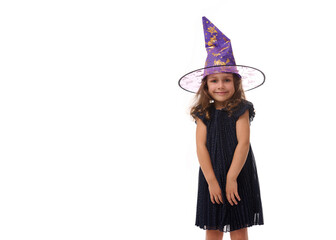 Portrait of pretty little girl wearing a wizard hat and dressed in stylish carnival dress, looking at camera posing with crossed arms against white background. Halloween concept with copy space