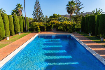 Views of a swimming pool with crystal clear waters surrounded by gardens, on a sunny morning. 