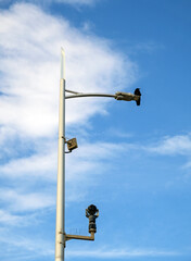 A crow perching on a lamppost and CCTV camera mount with a background of blue sky and clouds