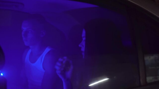 A young pleasant couple is smoking in the back seat of a car in a neon light. A man hovers in a vape and smokes. Everything is in smoke, I'm shooting a clip. overnight, parking.