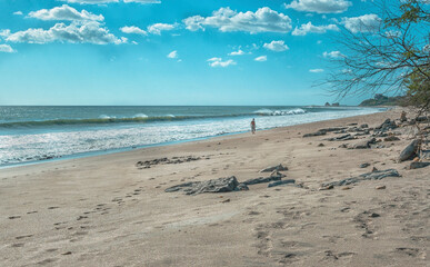 A man walking near the beach with waves on the shore and rocks in the background on a sunny day