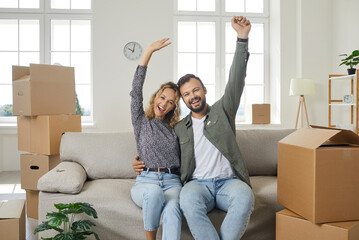 Portrait of happy excited young married couple sitting on sofa in new home, raising arms, looking...