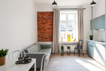 Small interior of loft apartment in industrail style with window and bricky wall. Cozy living room and kitchen with furniture, sofa and table with chairs.