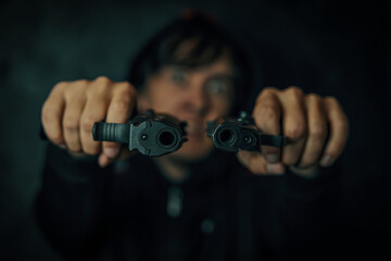 Murderer in hood on black background. Guy threatens with firearm. Two pistols in man's hands are pointed at camera. Close-up of two gun muzzles. Criminal with weapon.