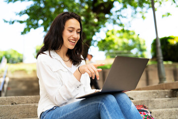 Young businesswoman sitting on steps outdoors and working on laptop. Beautiful girl learning in the park.