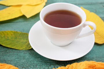 White cup of tea on wooden background among yellow fall leaves and knitted clothes