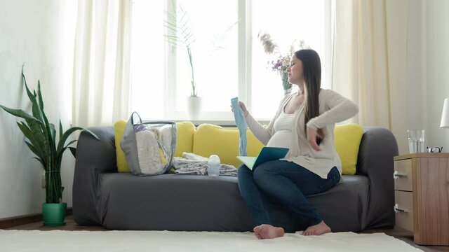Cute Pregnant Woman Writing Packing List For Maternity Hospital Notebook Prepares Bag. Young Ledy In Pregnancy Have Fun Spend Time On Yellow Gray Sofa At Home. Motherhood, Medicine Health Care Concept