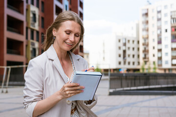 Business woman writing with pen in notebook. Caucasian successful girl in light jacket stands near an office building. Recording a daily routine