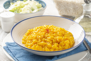 Italian dinner with risotto alla milanese and fresh salad. Italian dish made from saffron, rice, butter, hard cheese and vegetable broth. Raw arborio rice on background. White table. - obrazy, fototapety, plakaty