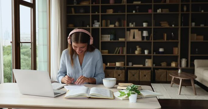 Laughing female student sit at home work desk in headset confer with teacher on virtual workshop in computer web app. Happy young teenage lady enjoy distant learning having fun studying subject online