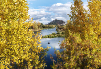 Autumn landscape with trees in the foreground. A river and a mountain can be seen between the yellow leaves. Cloudy sky.