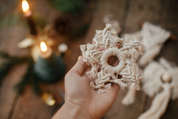 Hand holding stylish christmas macrame ornament on background of candles, fir branches on rustic...