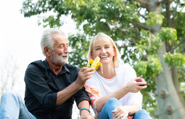 Fototapeta premium Senior caucasian couple sitting on field grass and enjoy eating banana, apple together outdoors in parks. Happy mature couple eating fruits in a park. Healthy and lifestyle retired couple outdoors.