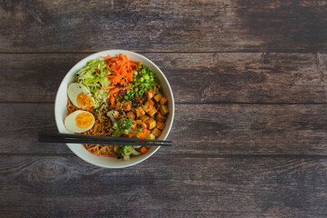 Variation of ramen homemade soup with noodles, marinated tofu, chopped carrots, onions, lettuce and broccoli in a bowl lying on a wooden table.