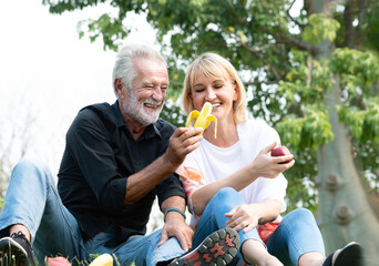 Senior caucasian couple sitting on field grass and enjoy eating banana, apple together outdoors in parks. Happy mature couple eating fruits in a park. Healthy and lifestyle retired couple outdoors.