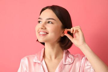 Young woman putting ear plug on pink background