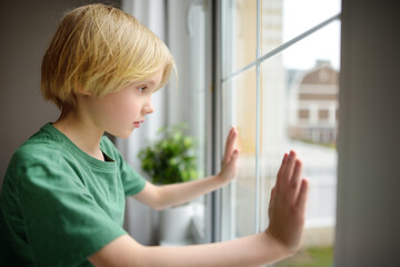 Sad little boy is sitting near window and watching street. Lonely at home. No friends, no siblings. One baby in family. Child cannot build relationships with his peers. Upset offended kid.