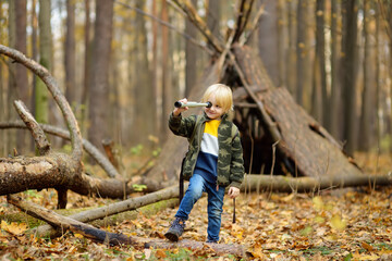 Little boy scout with spyglass during hiking in autumn forest. Child is looking through a spyglass. Behind the baby is teepee hut. Concepts of adventure, scouting and hiking tourism for kids.