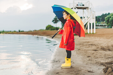 Boy in a red raincoat and yellow rubber boots stands at river bank and holding rainbow umbrella....