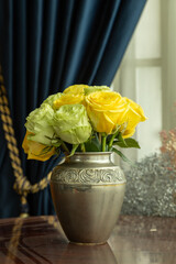metal vase with a bouquet of yellow and white roses on a wooden table with carving, decorative element in the interior of the home, in the background a curtain