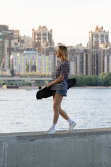 Adult freedom: woman in 40s walk near river holding longboard over big city view. Modern female enjoy skating after work wearing jeans shorts and casual t-shirt. Carefree skateboarder lady at sunset