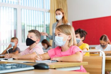 Portrait of schoolgirl in protective face mask who writing exercises at lesson in primary school