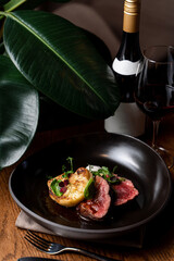 Advertisement photo of a wagyu steak on a black dish with a bottle of red wine, surrounded with green tropical leaves
