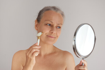 Smiling senior woman massaging her face with a jade roller while looking in the mirror © evafesenuk