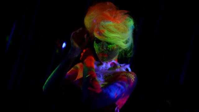 a woman with multicolored hair is dancing on a dark background. the skin is painted with bright glowing colors. the camera is moving