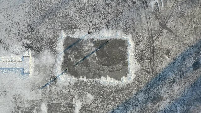 An aerial drone shot looking straight down on a makeshift ice rink on a large frozen lake with 3 people playing with a ball - Daytime. 