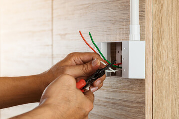 Close up hand of an electrician is stripping electrical wires in a plastic box on a wooden wall to...