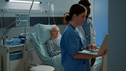 Multi ethnic hospital staff looking at medical tests on laptop for patient sitting in bed. Nurse and african american doctor consulting about healthcare treatment and injury recovery