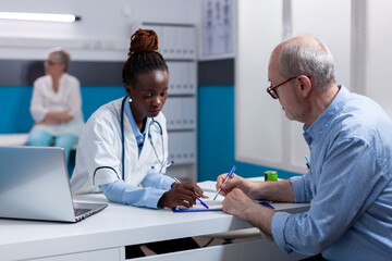 Obraz premium Black woman with medic profession looking at documents with elderly patient sitting at white desk. African american doctor and old man with disease writing on paper files for checkup