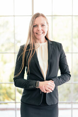 Confident Caucasian businesswoman wearing a suit, looking at the camera and smile in good mood, standing with blur office windows in the background. Vertical photo. Business Woman portrait.