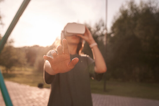 Close-up of a girl holding virtual reality glasses on her head with her hands, standing on a background of green trees. He holds out one hand to the camera. High quality photo