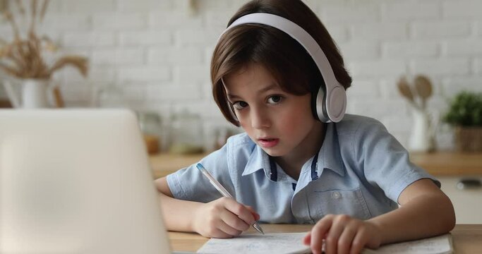 Concentrated little 7s kid boy wearing wireless headphones listening educational lecture in online school, attending virtual classroom lesson, communicating distantly with teacher, e-learning concept.