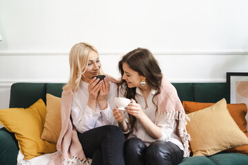 Obraz na płótnie Canvas Beautiful young woman and adult girl are drinking tea, talking and smiling while sitting on couch at modern home
