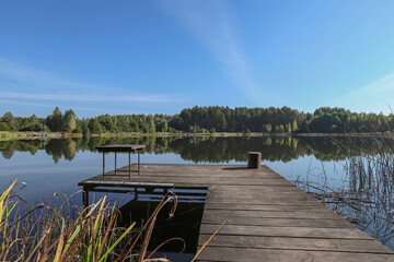 Landscape with wooden long jetty with chair for fishing, lake, forest on horizon and clear blue sky in summer.