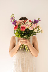 Colourful flowers bouquet in female hands on white background. Young woman in neutral beige washed linen dress hide  face. Aesthetic beauty and fashion floral concept. Face hidden by flower bouquet