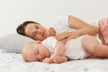 Fototapeta na wymiar Happy pleased young dark haired mother and her baby lying together on bed, mommy hugging her charming daughter, people wearing white clothing, motherhood and childhood.