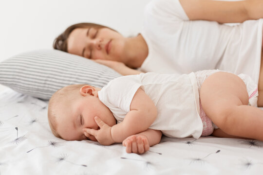 Side view of of young adult mother and her baby sleeping together lying on bed, posing at home in light room, mommy can have a rest while daughter sleeps.