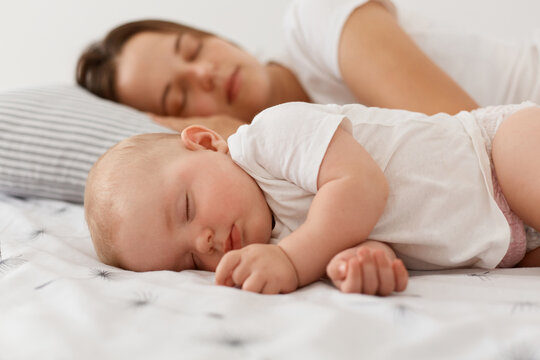 Portrait of cute charming baby girl wearing white clothing lying and sleeping near her lovely mother on bed, having nap, resting at home near mommy with closed eyes.