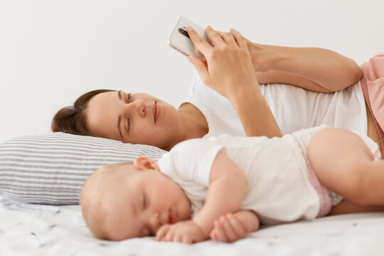 Portrait of sleeping toddler daughter wearing white attire lying near mother, woman using smart phone when wakes up and checking emails or social networks.