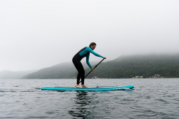 A woman in a wetsuit paddle a paddleboard with an oar on the sea waves in cloudy weather.
