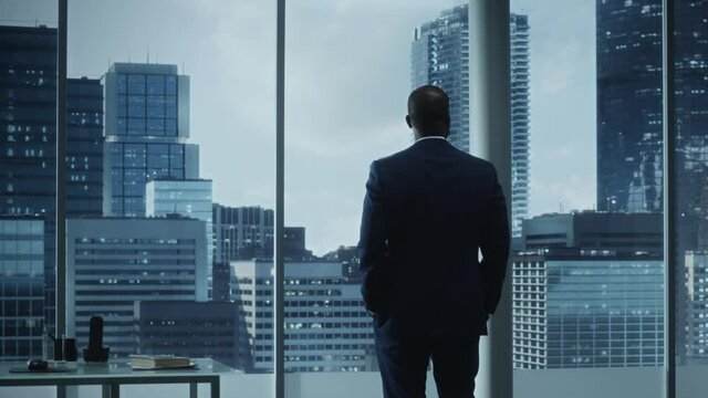 Thoughtful African-American Businessman in a Perfect Tailored Suit Standing in His Office Looking out of the Window on Big City. Successful Investment Manager Planning Strategy for e-Commerce Startup