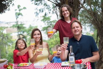 Portrait of happy smiling family at picnic. Mid adult parents sitting around table with sons, cheering at camera. Family time, outdoor activity, social media concept