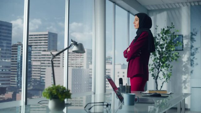 Successful Muslim Businesswoman Wearing Suit and Hijab Standing in Office Looking out of Window on Big City. Confident Female Digital Entrepreneur Planning Investment Strategy for e-Commerce Startup